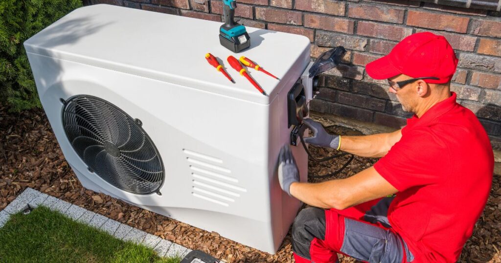 A man in a red shirt and cap, performing safety checks on a large heat pump outdoors.